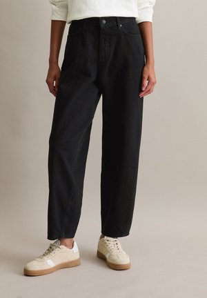 Person med løse sorte jeans og beige sneakers står mot en ensfarget beige bakgrunn.