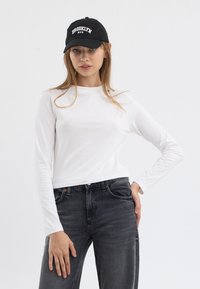 Hvit langermet topp med rund hals, parret med mørkegrå jeans. Svart caps med "BROOKLYN NYC" i hvit tekst.