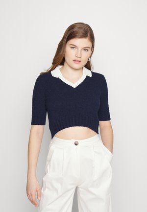 Sweater cortado em azul-marinho com uma textura suave, mangas curtas e um colarinho branco. Apresenta um decote em V e bainha canelada. Usado com calças brancas de cintura alta.