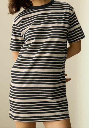 Vrouw die een zwart-wit gestreepte jurk met korte mouwen draagt, staand met handen achter de rug tegen een effen achtergrond.