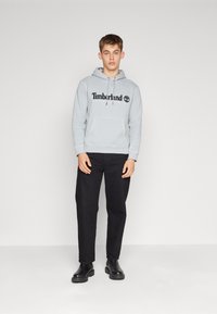Felpa grigia chiara con logo Timberland nero, tasca a marsupio frontale, abbinata a pantaloni neri e stivaletti neri, in piedi su uno sfondo neutro.