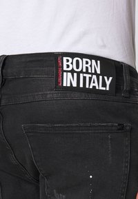 Los pantalones vaqueros de mezclilla negra presentan un acabado texturizado con un patrón desgastado. Una etiqueta prominente dice "NACIDO EN ITALIA", con detalles en rojo y blanco.