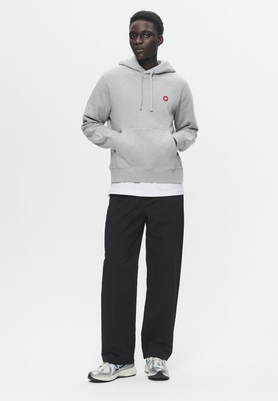 Grauer Hoodie mit rotem Logo, Kängurutasche und verstellbarem Zugverschluss an der Kapuze, kombiniert mit schwarzen Hosen und silbernen Sportschuhen.