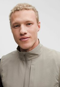 Světle šedý zip-up vest s texturovaným povrchem, stojáčkem a elegantním designem, nošený přes černou dlouhou rukávovou košili.