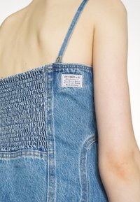 Personne portant une robe en jean bleu avec un dos élastiqué froncé et une étiquette Levi Strauss & Co. sur le côté droit près de la bretelle de l'épaule.