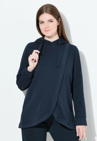 Donna che indossa un top a maniche lunghe con cappuccio di colore blu navy, con un pannello frontale sovrapposto, in piedi mentre tiene un cordoncino vicino al collo.