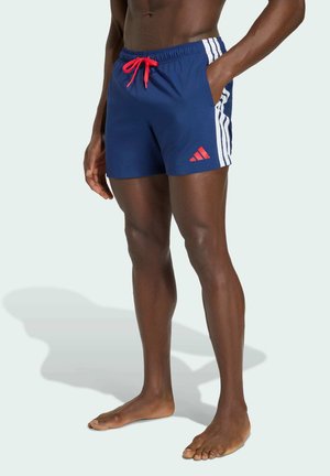 Persona descalza que lleva pantalones deportivos azul marino con rayas blancas en los costados, cordón rojo y logo rojo de Adidas en la pierna izquierda.