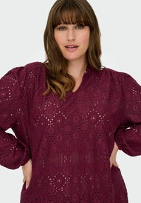 Burgundska bluzica z teksturirano zasnovo, podobno čipki, z diamanti in napihnjenimi rokavi. O-vrat in ohlapen kroj.