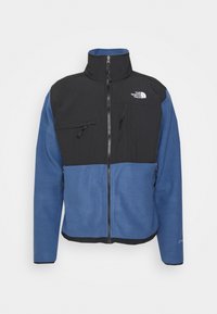 Kurtka polarowa z czarnym górnym panelem i niebieską dolną częścią, zapinana na zamek, z kieszeniami na zamek, wysokim kołnierzem oraz logo The North Face.