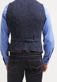 Mörkblå stickad väst med en figurnära design, med ett bälte med spänne på baksidan, ihop med en ljusblå långärmad skjorta.