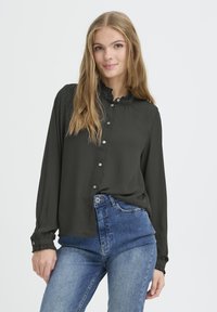 Dunkelgrüne Bluse mit langen Ärmeln, Rüschkragen, Knopfleiste und geknöpften Ärmeln, kombiniert mit hochtaillierten blauen Jeans.