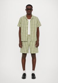 Zeleno-biele kostkované tričko s krátkym rukávom a zodpovedajúce šortky, vyrobené z ľahkej tkaniny. Čierne slnečné okuliare dotvárajú outfit.