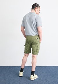 Tricou gri cu mânecă scurtă și pantaloni scurți cargo de culoare verde măslin, cu multe buzunare, văzuți din spate. Încălțămintea este o pereche de adidași de culoare deschisă.