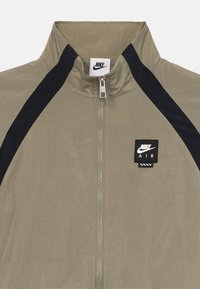 Αθλητικό μπουφάν Nike με φερμουάρ μπροστά, ψηλό γιακά, ελαφρύ ύφασμα σε ελαιόλαδο πράσινο χρώμα, μαύρες λεπτομέρειες στα πλάγια και τετράγωνη ετικέτα λογότυπου.