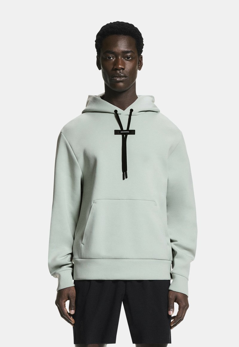 Mladić odjeven u svijetlosivu majicu s kapuljačom s crnim vrpcama i crne kratke hlače, stoji nasuprot jednostavnoj bijeloj pozadini.