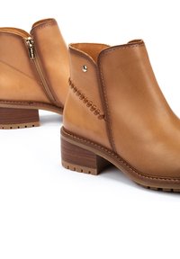 Bottes de cheville en cuir tan avec une fermeture éclair sur le côté, un talon en bois empilé et des détails de couture décoratifs sur le côté. Texture lisse et design structuré.