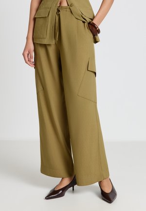 Femme portant un pantalon cargo ample olive avec des poches, associé à des talons pointus marron et un gilet olive assorti.