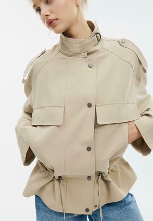 Femme portant une veste beige boutonnée avec col montant, poches à rabat et cordons ajustables à la taille, associée à un jean bleu.