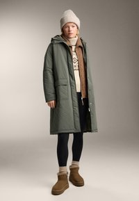 Geacă cu puf verde, matlasată, cu glugă, purtată cu un pulover crem cu model, colanți negri și botine maro până la gleznă. Căciulă tricotată bej pe cap.