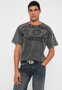 Muž v vybledlém černém tričku s potiskem a tmavě modrých džínách s černým páskem, stojící před jednobarevným světlým pozadím.