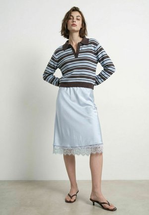 Femme portant un chemisier à manches longues rayé marron et bleu, une jupe bleu clair bordée de dentelle et des sandales noires à talons, debout, mains sur les hanches.