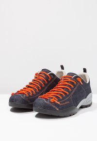 Farkkukengät kiipeilyyn, joissa on oranssit nauhat, harmaat kumipohjat ja vahvistetut kärjet, matala malli ja tekstiilinen päällinen.