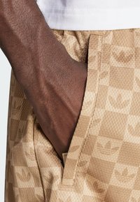 Beige shorts with a textured, checkered pattern featuring subtle Adidas logos. The fabric appears stretchy, with side pockets.