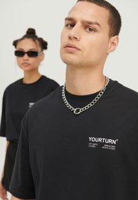 Bărbat tânăr purtând un tricou negru cu inscripția "YOURTURN" și un lanț argintiu la gât, femeie cu tricou negru și ochelari de soare, blurată în fundal.