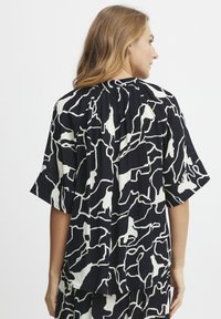 Sort og hvid mønstrede bluse med korte ærmer, der har abstrakte designs og en samlet halslinning. Lavet af letvægtsstof.