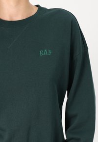 Sweat-shirt en coton vert foncé avec col rond large, poignets côtelés et logo "GAP" brodé dans une teinte de vert légèrement plus claire.