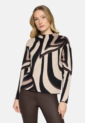Femme aux cheveux blonds portant un pull à manches longues à motifs abstraits beige, noir et marron, ainsi qu'un pantalon marron foncé ajusté, posant devant un fond blanc.