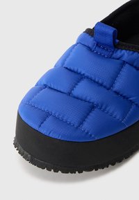 Kék steppelt papucs, párnázott felső résszel, texturált fekete gumitalppal és egy kis kék textil húzófüllel. Strukturált és könnyű kivitel.