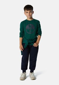 Camisa verde de manga larga con un logo circular, pantalones jogger en azul marino y zapatillas beige. La camisa tiene un detalle de cintura anudada.