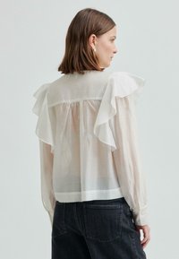 Kobieta z krótkimi brązowymi włosami, ubrana w przezroczystą białą bluzkę z falbaniastymi ramionami i długimi rękawami, zestawioną z ciemnymi dżinsami o wysokim stanie.