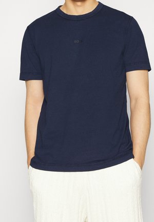 Person trägt ein schlichtes dunkel marineblaues kurzärmeliges T-Shirt mit dezentem Text auf der Brust und strukturierte weiße Hose, Hände in den Taschen.