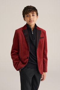 Veston en daim rouge avec revers et accents de poche en satin noir, porté sur un pull noir à col zippé et associé à un pantalon noir.