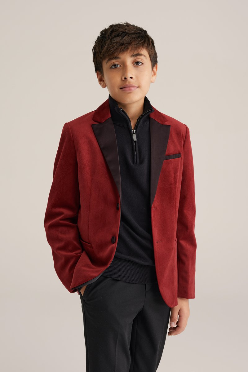 Veston en daim rouge avec revers et accents de poche en satin noir, porté sur un pull noir à col zippé et associé à un pantalon noir.