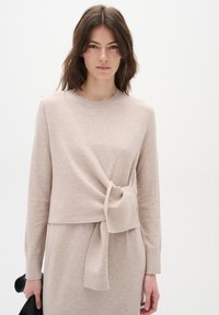 Světle béžový svetr s designem do oblouku, který má detail uzlu vpředu, dlouhé rukávy a jemnou texturu. Vhodný k sladěným šatům.