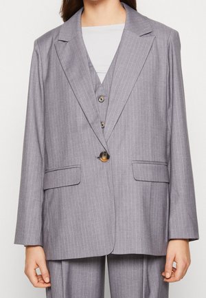 Mujer con chaqueta de traje gris de rayas diplomáticas, con chaleco y pantalones a juego, sobre una blusa blanca, abotonada en la cintura.