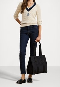 Krämfärgad kabelstickad tröja med marinblå V-ringning och logotyp, mörka jeans, svart tote-väska och svarta loafers. Fokusera på en enkel, avslappnad design.