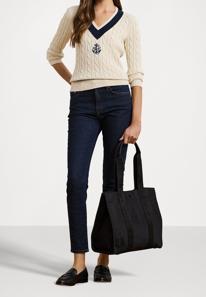 Krämfärgad kabelstickad tröja med marinblå V-ringning och logotyp, mörka jeans, svart tote-väska och svarta loafers. Fokusera på en enkel, avslappnad design.