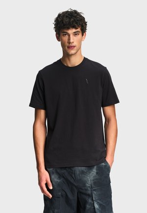 Giovane uomo con capelli ricci che indossa una maglietta nera a maniche corte semplice e pantaloncini cargo scuri, in piedi davanti a uno sfondo grigio chiaro.