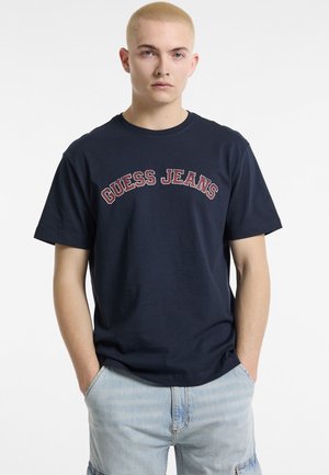 Tumši zili kokvilnas T-krekli ar "GUESS JEANS" logo sarkans krāsā, kas izvietots uz krūtīm. Klasisks apaļais kakls, īsas piedurknes un brīvs piegriezums.