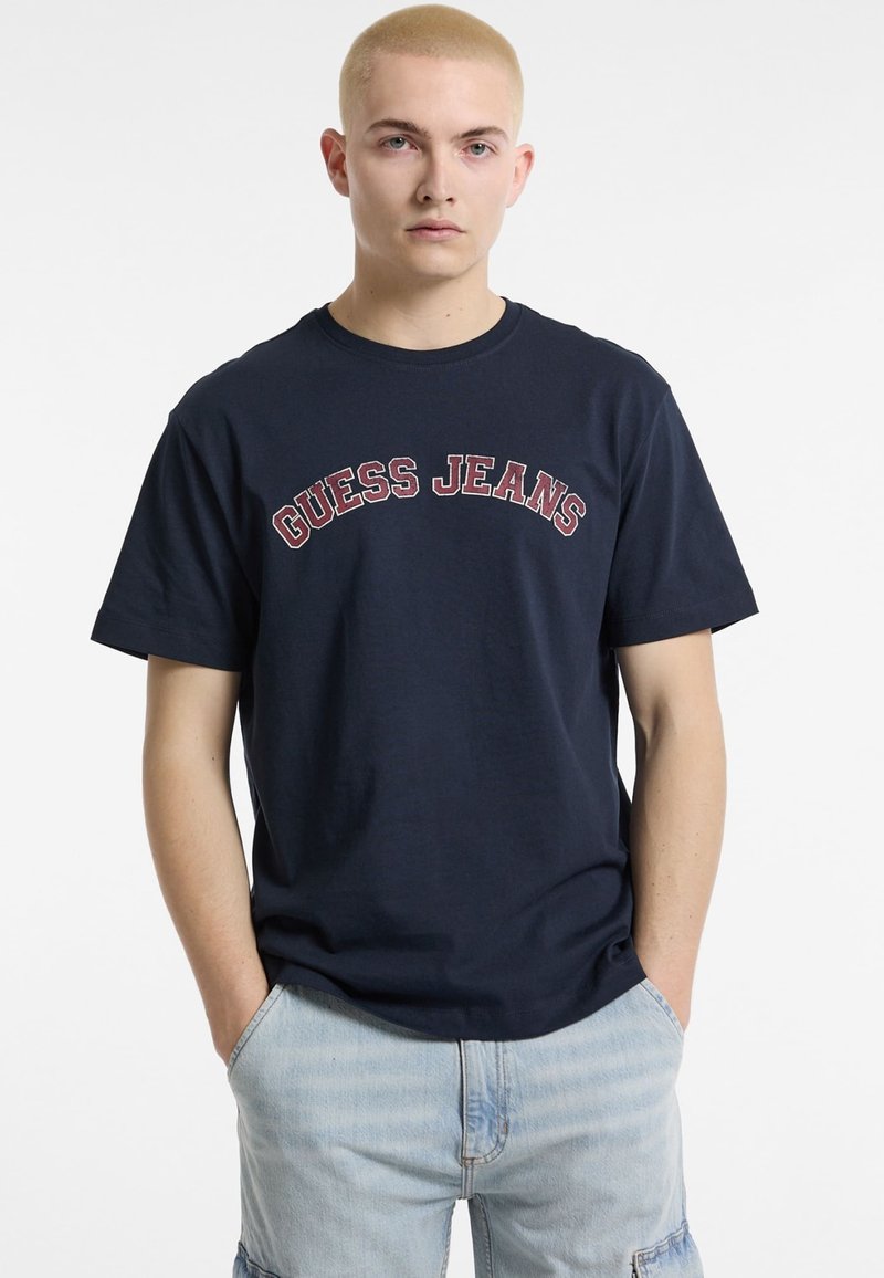 Mornaroplava pamučna majica kratkih rukava s "GUESS JEANS" logom u crvenoj boji preko grudi. Klasičan izrez na vratu, kratki rukavi i opušteni kroj.