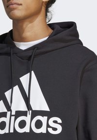 Sweat à capuche noir avec un grand logo Adidas blanc. Doté d'une capuche avec cordon de serrage et d'un tissu texturé. Couture visible et ouverture de cou large.