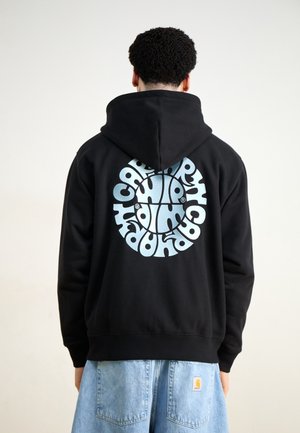 Person with short curly hair wearing black hoodie with blue circular graphic and light blue jeans, facing away from the camera.