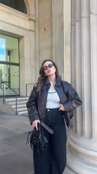 Braune Lederjacke mit hohem Kragen, grauem Tanktop, hoch taillierten schwarzen Jeans und einer kleinen schwarzen Handtasche mit Fransen.