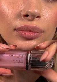 3D Hydra Xtreme Lipgloss i en rosa genomskinlig flaska med ett silverlock, hålls nära fylliga läppar med en ljusrosa nyans och en glansig yta.