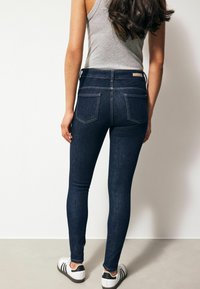 Jeans de mezclilla oscura ajustados con una cintura de altura media, dos bolsillos traseros y costuras en contraste, combinados con zapatillas deportivas blancas.