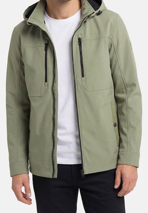 Mand iført en lysegrøn hættejakke med sorte lynlåse over en hvid T-shirt og sorte bukser, stående mod en ensfarvet baggrund.